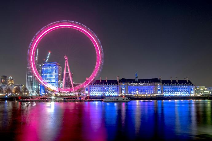 Londra’da gece çekilmiş bir manzarada pembe ışıklarla aydınlatılmış London Eye dönme dolabı ve Thames Nehri kıyısındaki tarihi bina görülüyor; nehir yüzeyinde renkli ışıkların yansımaları dikkat çekiyor.